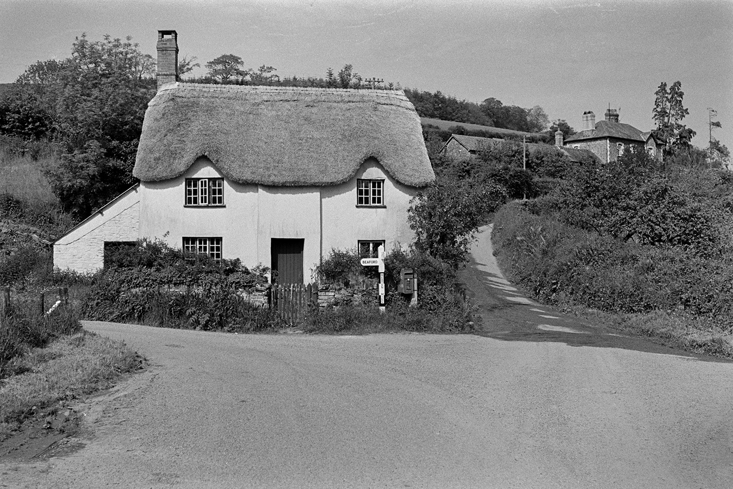 A thatched cottage, with eyebrow eaves, at a road junction at Merton Mill near Merton. A signpost and post box are visible by the wall outside the cottage.