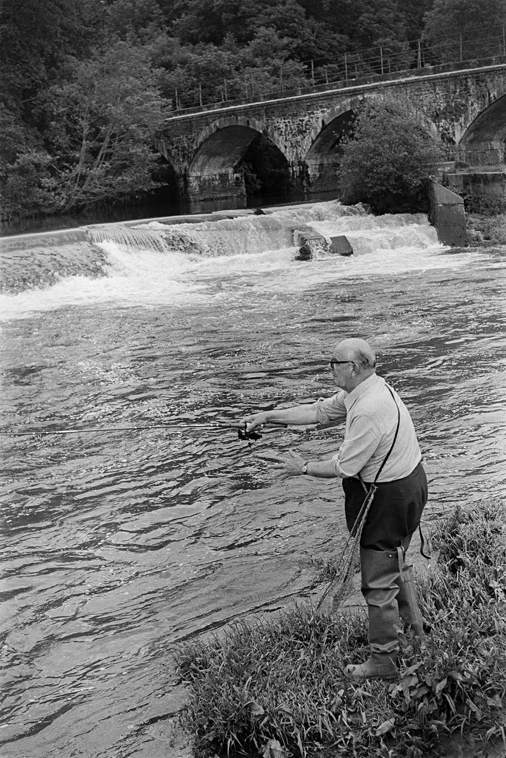 Dr J V Morris, OBE, fishing in the River Torridge on a fishing holiday at the Half Moon Inn, Sheepwash. He is wearing waders and is using a fishing rod. A weir and bridge over the river are visible in the background.