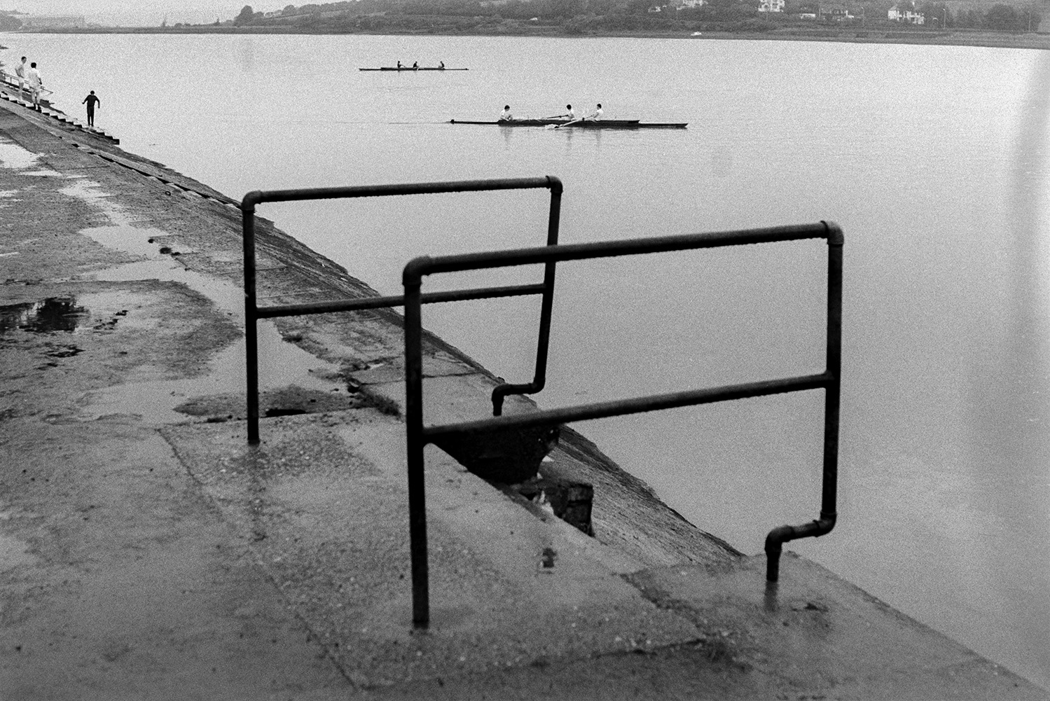 Railings by steps down a concrete bank to the River Torridge at Bideford. Rowers can be seen on the river and North Gate is visible on the other side of the river. The railings were later removed.