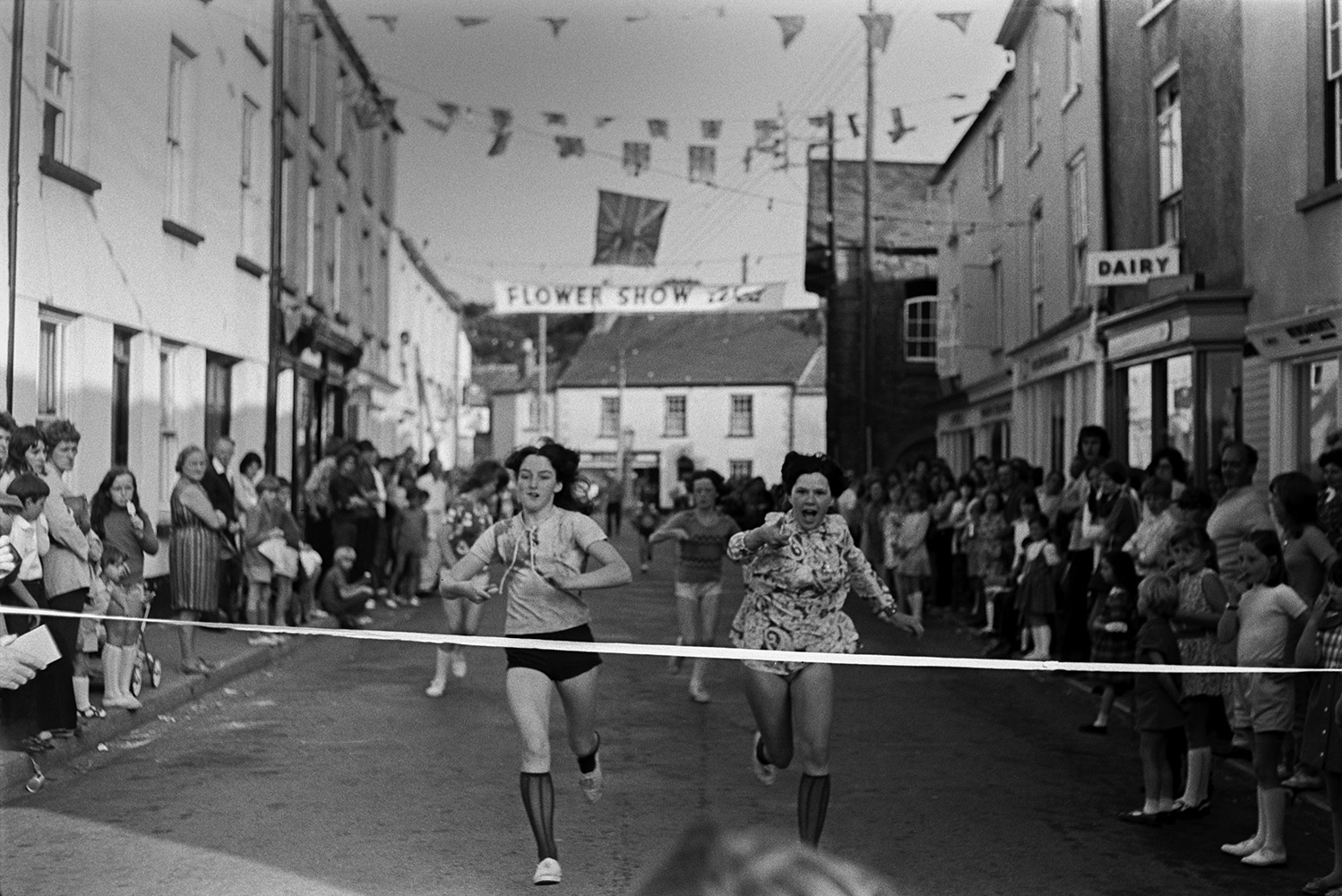 Girls crossing the finishing line of a race in  street at Chulmleigh Fair. Spectators are watching from the sides of the road and the street is decorated with bunting.