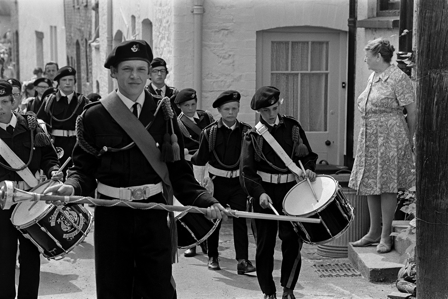 A woman watching a boys drum band parading through a street at Chulmleigh Fair. The boy leading the band is carrying a large mace.