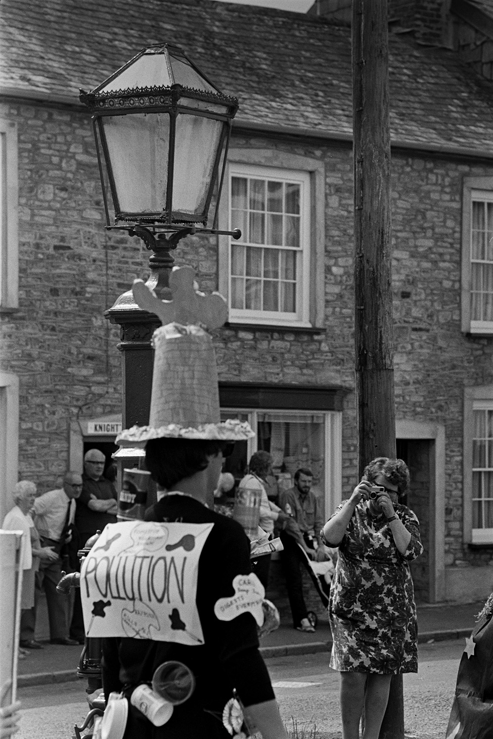 A woman taking a photographs of a person in fancy dress by a lamp in a street at Chulmleigh Fair. They are wearing a costume titled 'Pollution' with various bits of rubbish stuck to their clothes.