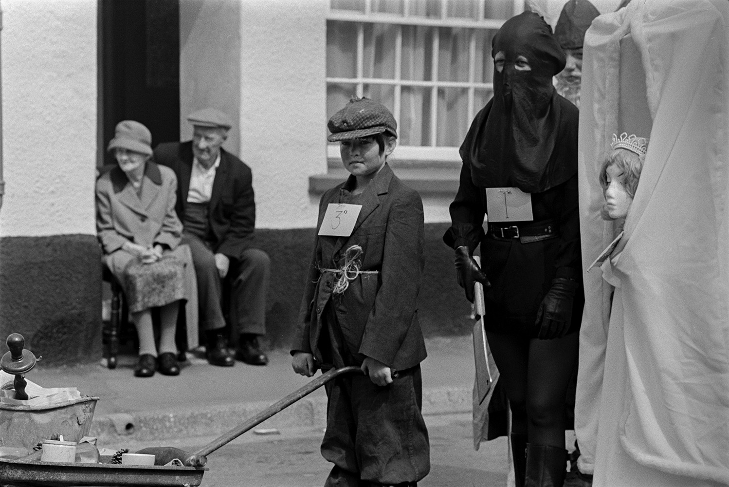 A man and woman sat in their doorway watching children in fancy dress parading through the street at Chulmleigh Fair. One child is dressed as an executioner.