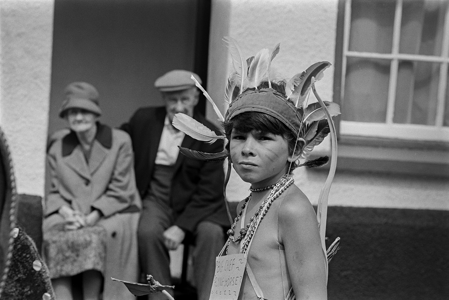 A man and woman sat in their doorway watching children in fancy dress parading through the street at Chulmleigh Fair. One child is dressed as an Indian Chief.