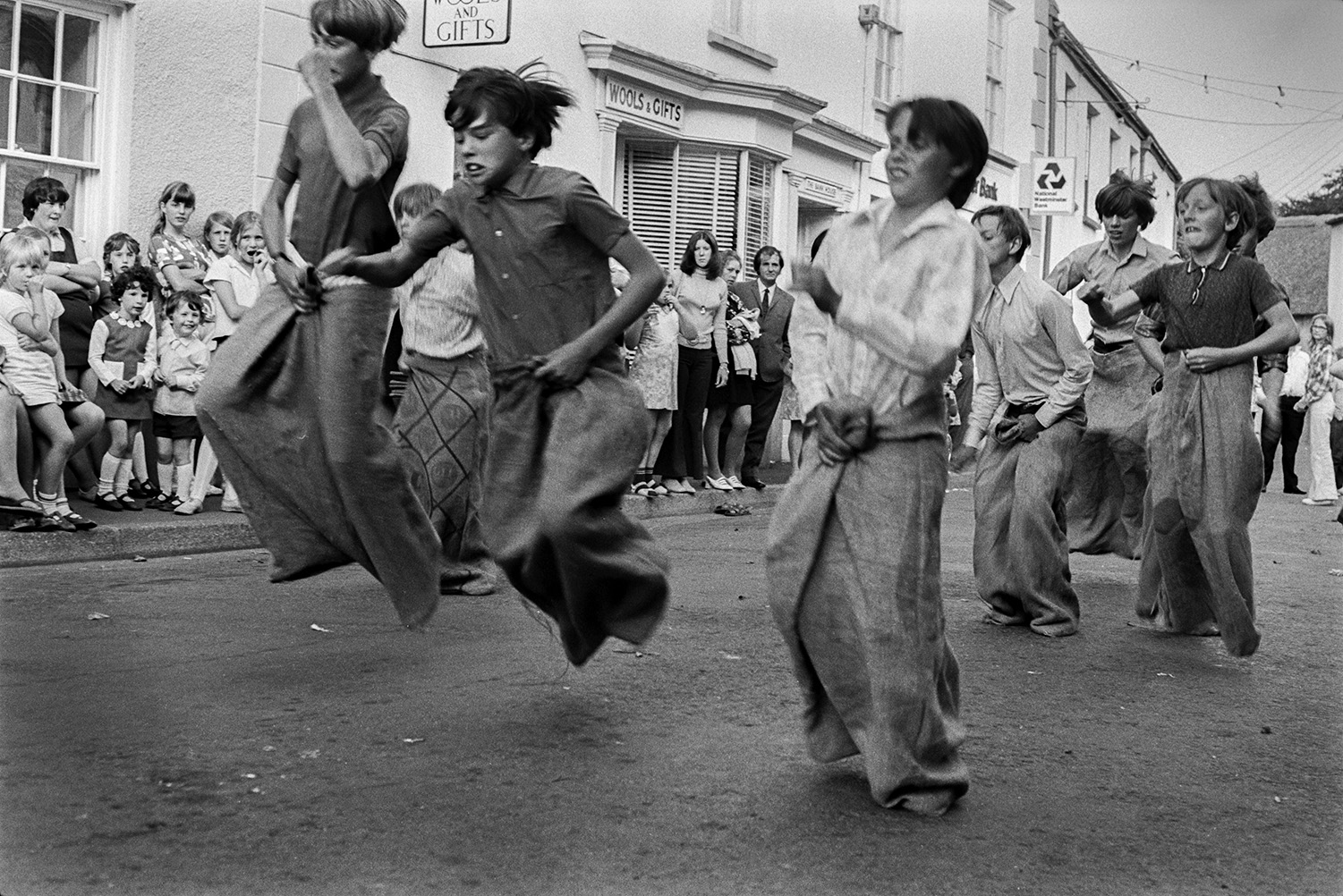 Boys competing in a sack race through a street at Chulmleigh Fair. Spectators are watching outside the shop front of 'Wools & Gifts'.