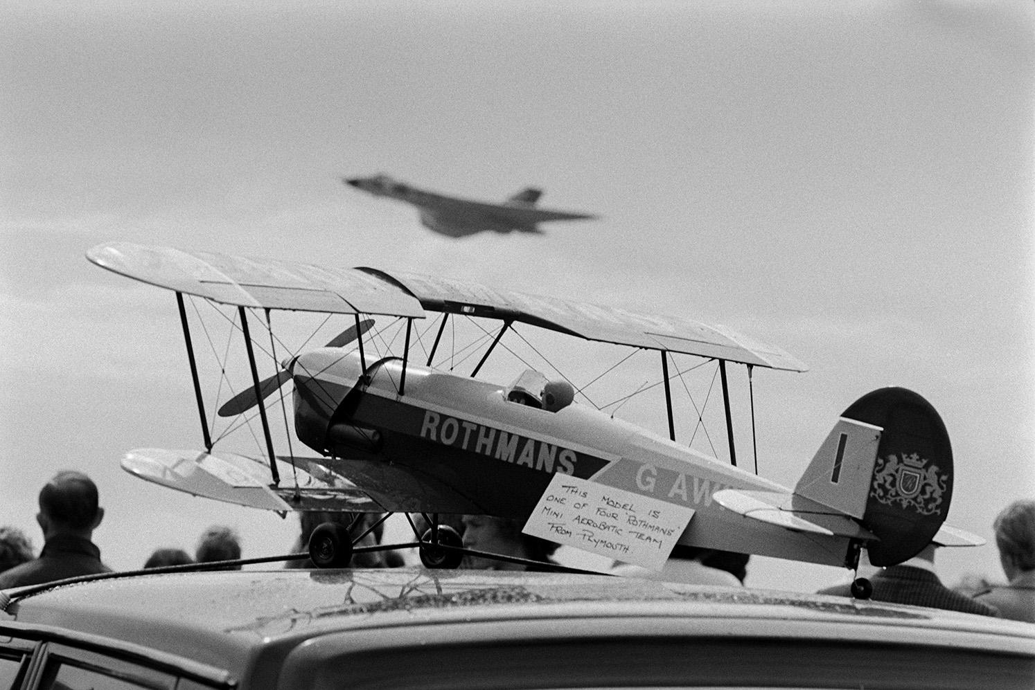 Spectators watching an aeroplane at the Chivenor Air Show, at the Royal Marine Base in Braunton. In the foreground a model of a Rothmans plane is visible. A sign next to it states it is one of four Rothmans Mini Aerobatic team from Plymouth.