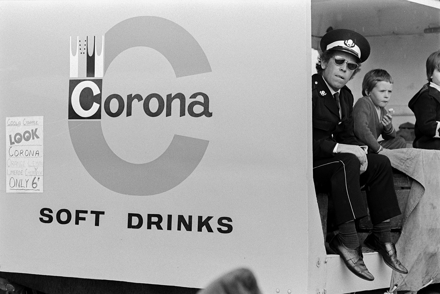 A Corona soft drinks advert on the side of a viewing booth at the Chivenor Air Show at the Royal Marine Base in Braunton. A man in uniform, possibly a pilot, is sat inside the booth with two children.