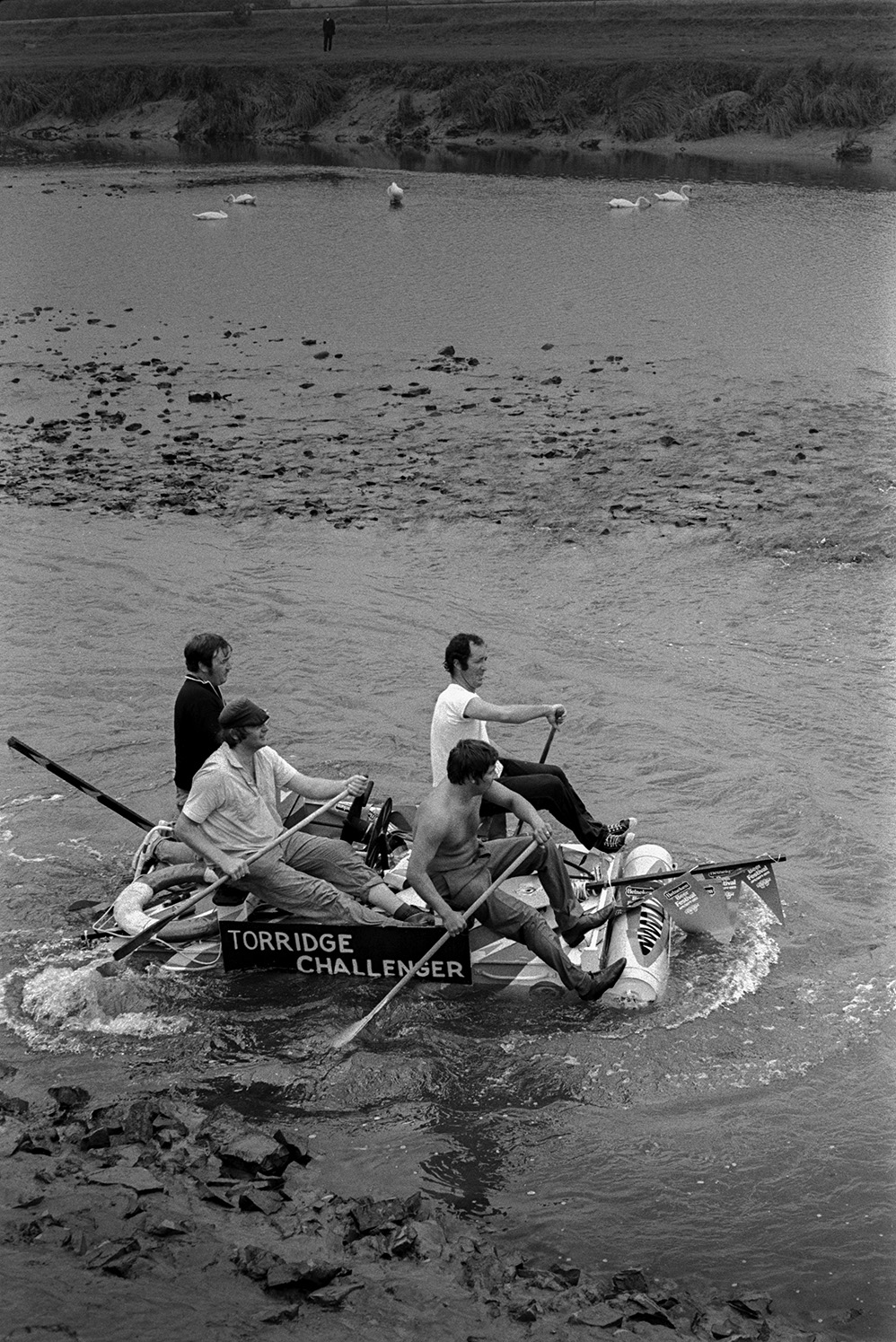 Four men rowing a homemade raft called the 'Torridge Challenger' in the Barnstaple Raft Race in Rock Park, Barnstaple. Swans can be seen on the water in the background.