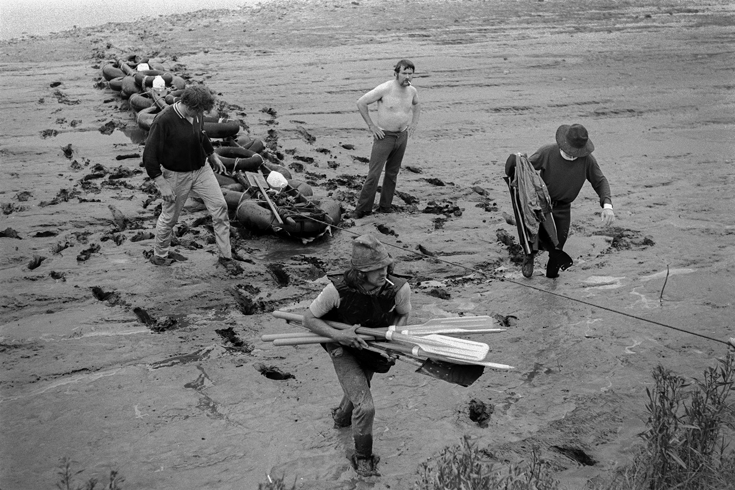 A homemade raft being dragged across mud flats after the Barnstaple Raft Race in Rock Park, Barnstaple. Four men who rowed the raft are walking through the mud alongside it. One of them is holding the oars.