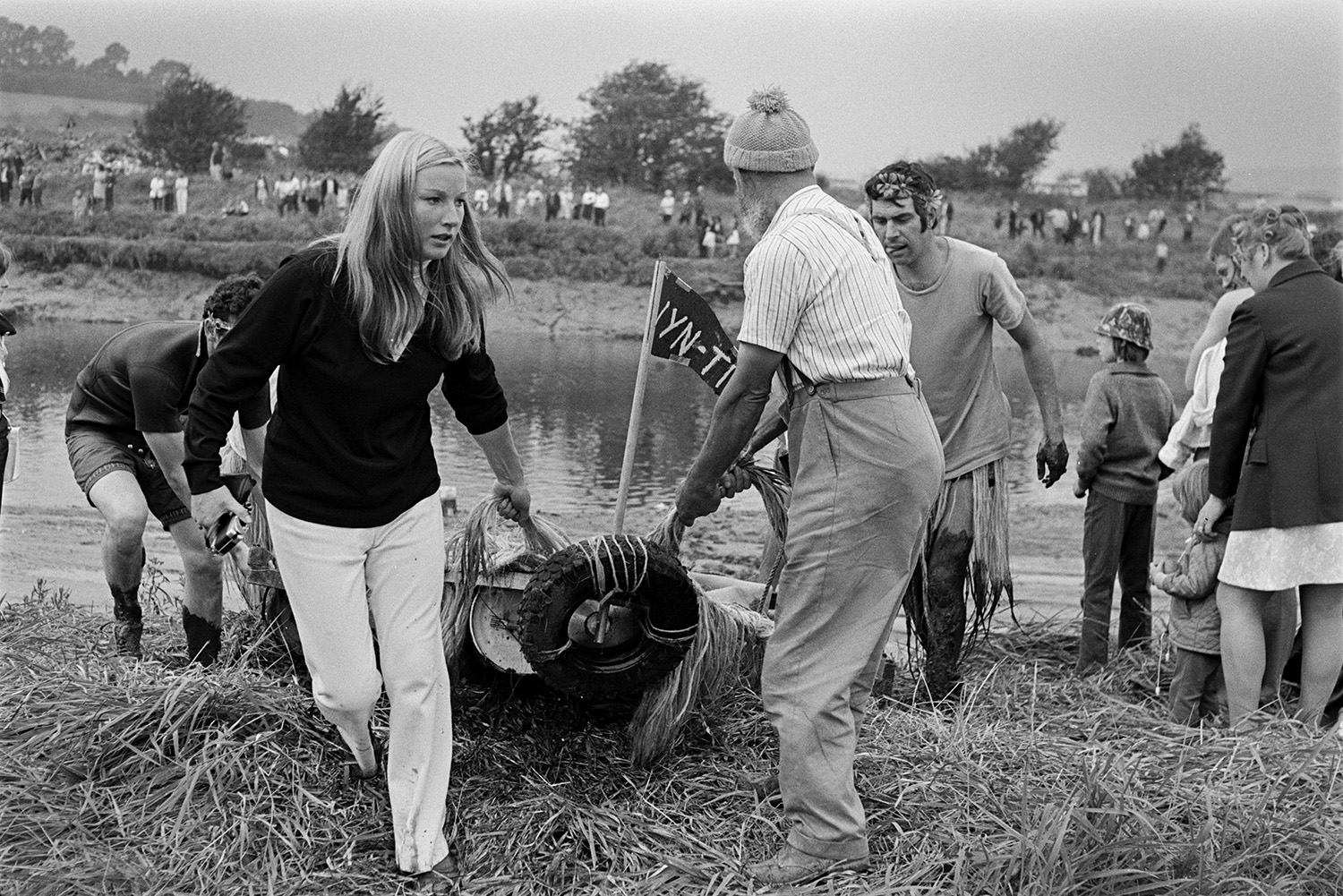 A woman and men dragging a homemade raft out of the water at Barnstaple Raft Race in Rock Park, Barnstaple. Spectators are watching race on the opposite side of the river.