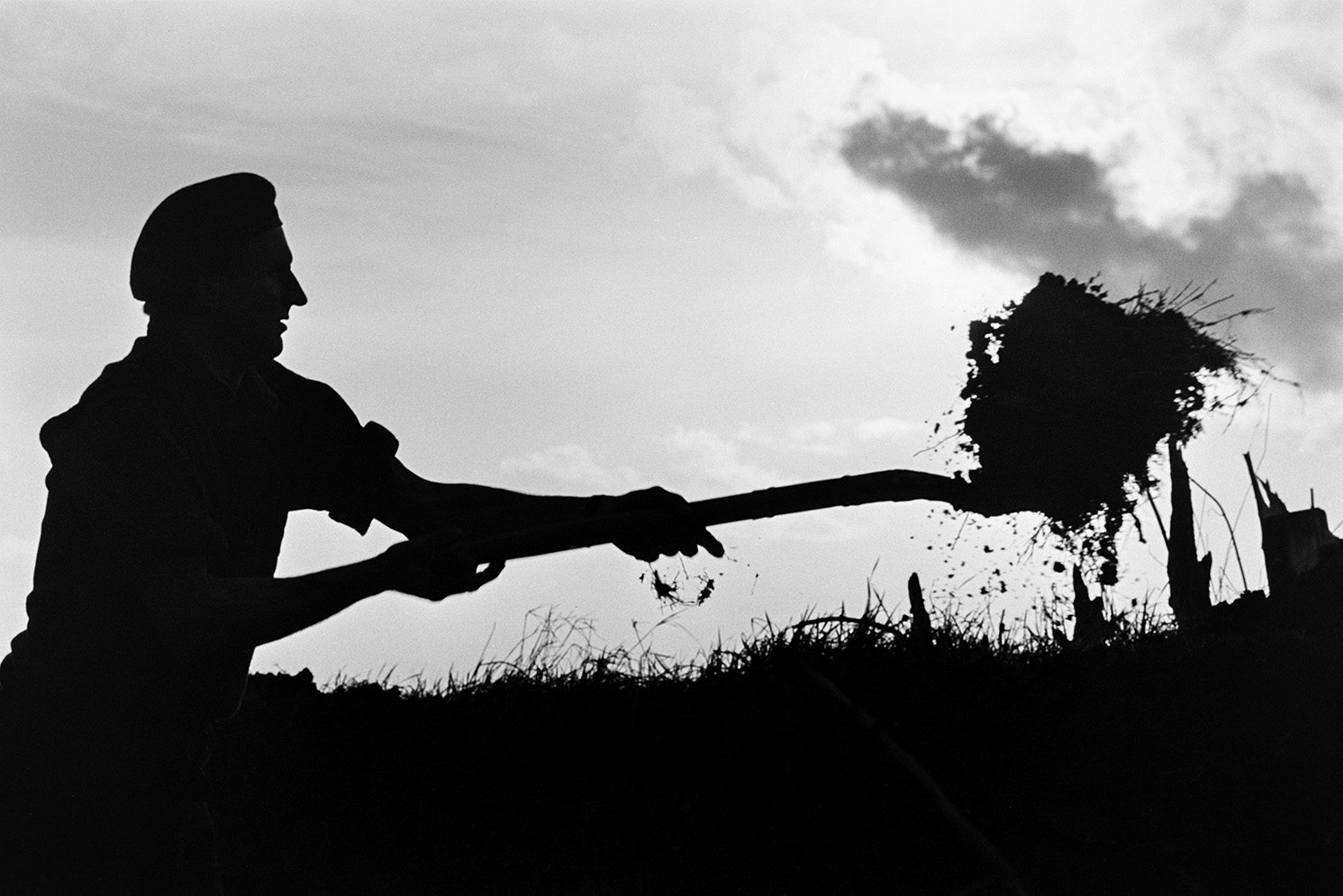 Ivor Bourne clatting a hedge at Mill Road Farm, Beaford using a fork. He is building up hedge bank with turf and is silhouetted against the horizon. The farm was also known as Jeffrys.