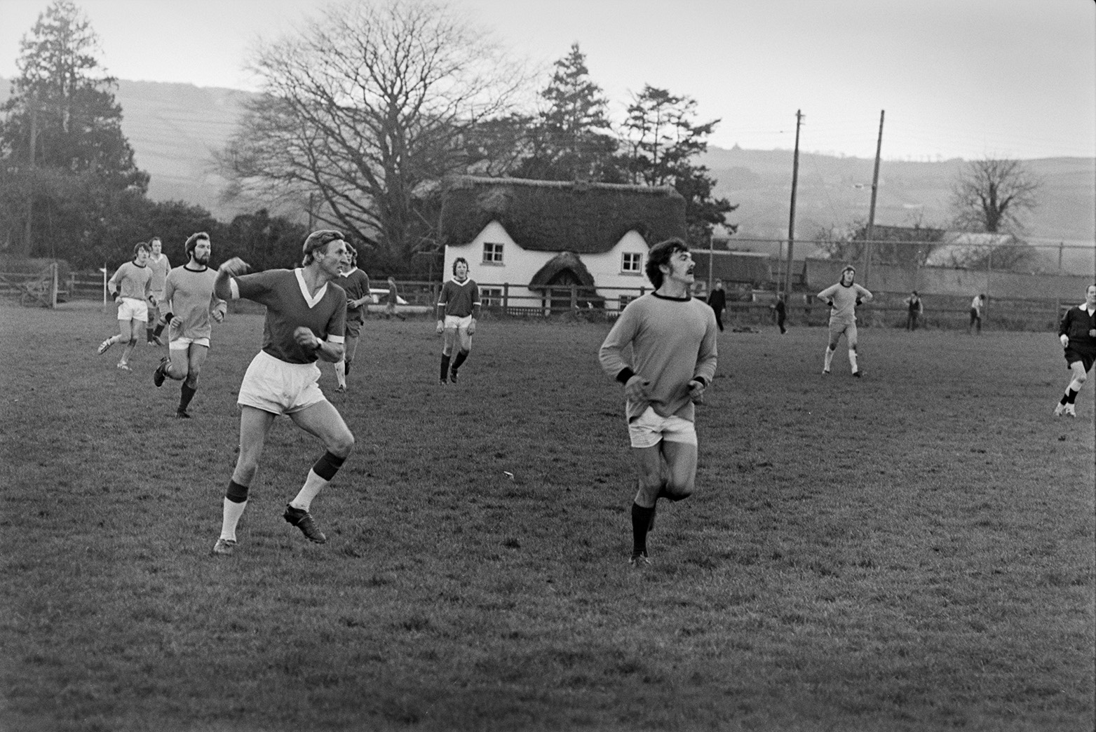 Football match between Merton and Dolton at Merton. Football players are on the pitch. Ivor Bourne is in the foreground on the left and John Kelway is on the right. The player between them further back is possibly John Woodland and the man with his hands on his hips is possibly Les Jury. The thatched cottage in the background is New Road Cottage.