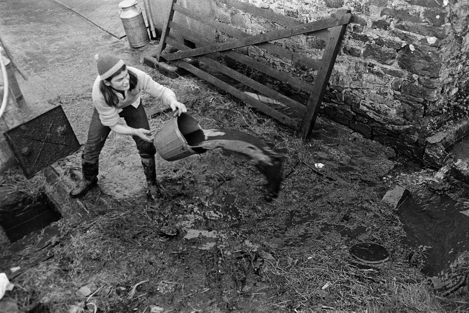 Derek Bright emptying a bucket in the farmyard at Mill Road Farm, Beaford. He is stood next to an open manhole or drain. A wooden field gate is lent by a milk churn and a stone building in the background. The farm was also known as Jeffrys.