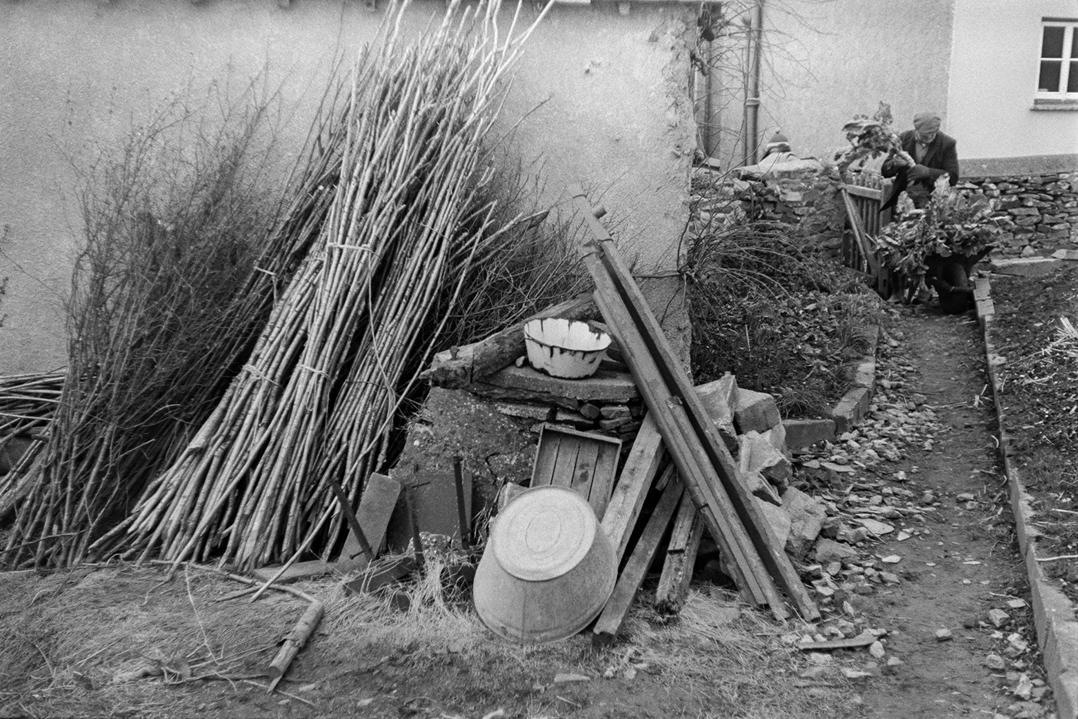 Various items, including bean sticks, a tin bath, planks of wood and branches stacked against the wall of a house, in Beaford. In the background a man is walking through a garden gateway with a wheelbarrow full of vegetables.