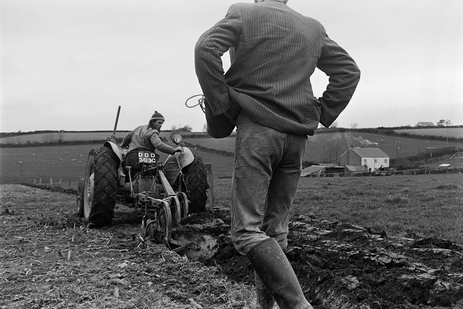 Derek Bright ploughing a field so it is ready to plant corn at Mill Road Farm, Beaford. He is looking over his shoulder to check the furrow. A man is walking along the field, following him and a cottage can be seen in the background. The farm was also known as Jeffrys.