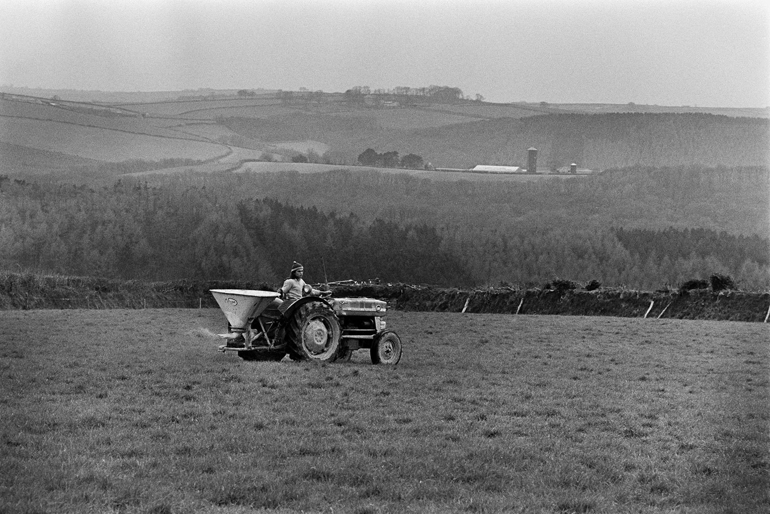 Derek Bright spreading fertilizer on a field at Mill Road Farm, Beaford using a tractor and fertilizer spreader. A landscape of fields, woodland and hedgerows can be seen in the background. The farm was also known as Jeffrys.