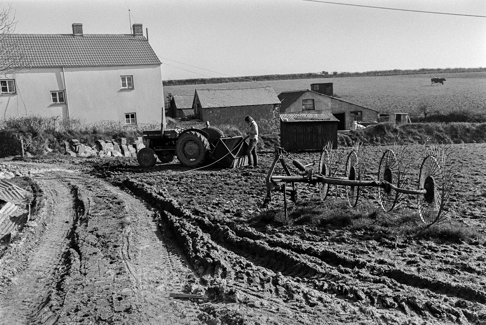 Derek Bright washing out a link box with a hose in a muddy field at Mill Road Farm, Beaford.  A whisk is lying next to a muddy track in the foreground and a farmhouse and barns can be seen in the background. The farm was also known as Jeffrys.