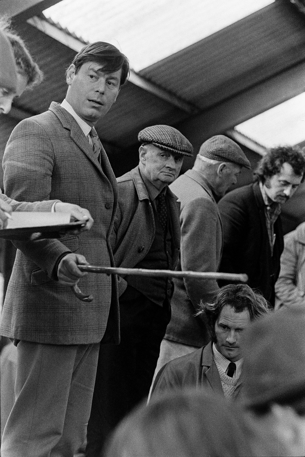 Hatherleigh Market. Men stood on boards above sheep pens and looking at the livestock below. The man holding a walking stick is possibly the auctioneer.