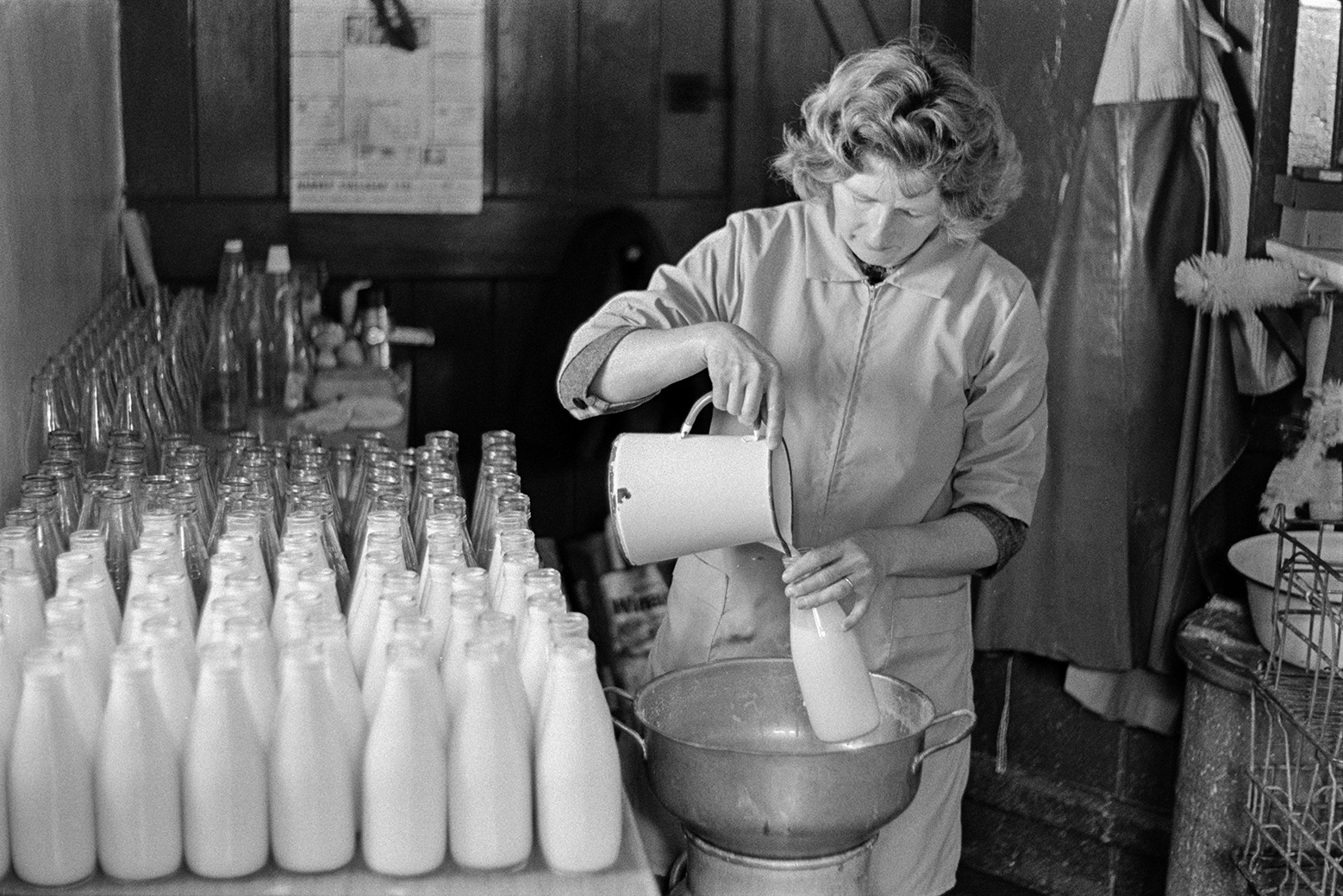 A woman filling milk bottles with milk in a dairy at Mill Road Farm, Beaford. She is filling the bottles over a ilk churn. The farm was also known as Jeffrys.