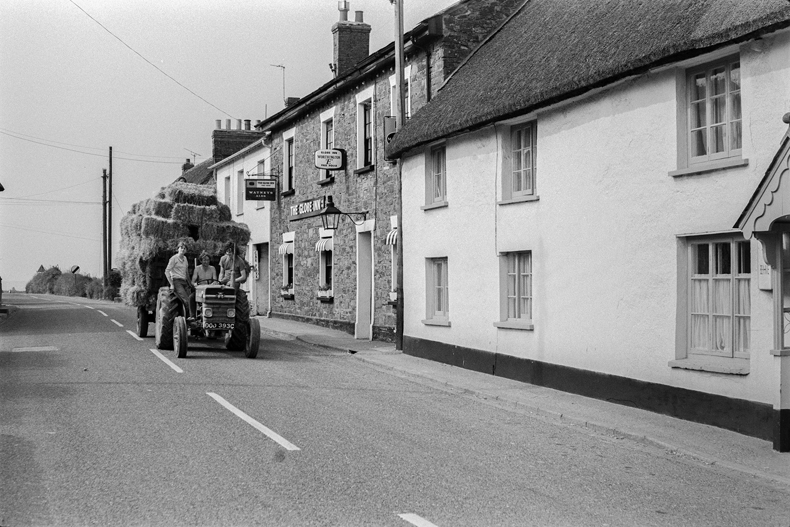 Two men and a woman transporting hay bales with a tractor and trailer down a street in Beaford, past The Globe Inn. They are all sat on the tractor. Ivor Bourne is driving the tractor.
