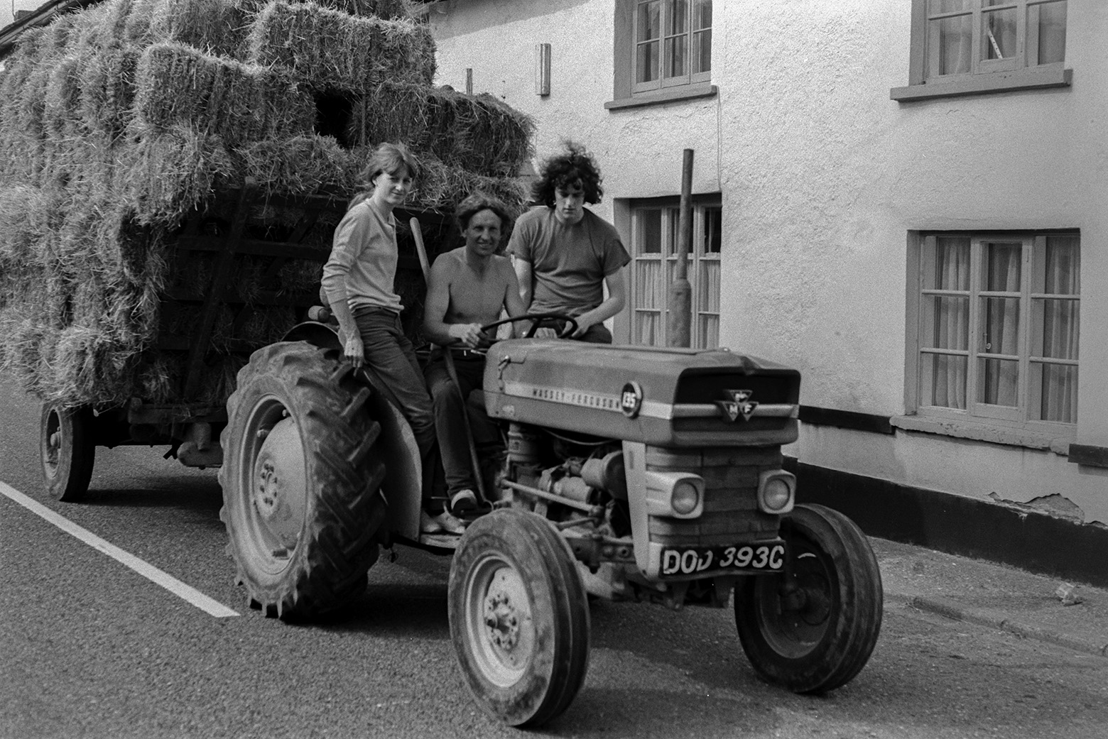 Two men and a woman transporting hay bales with a tractor and trailer down a street in Beaford. They are all sat on the tractor. Ivor Bourne is driving the tractor.
