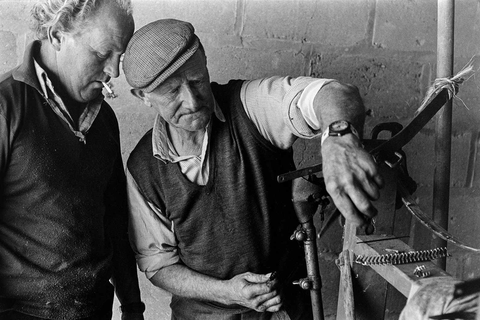 Two men looking at shearing machine in a barn at Mill Road Farm, Beaford. One of the men is smoking a cigarette. The farm was also known as Jeffrys.