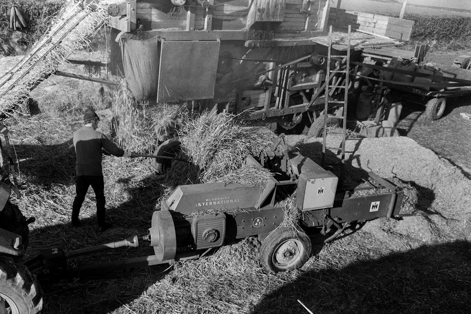 A baler next to a reed comber in a field at Beaford. Reed can be seen coming off the reed comber. A man is stacking some of the reed next to a baler in the foreground.