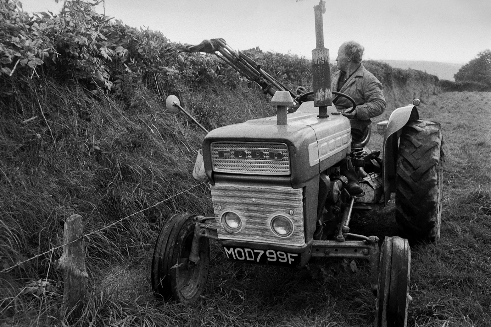 A man cutting a hedge along the edge of a field in Beaford, using a tractor and hedge trimmer.