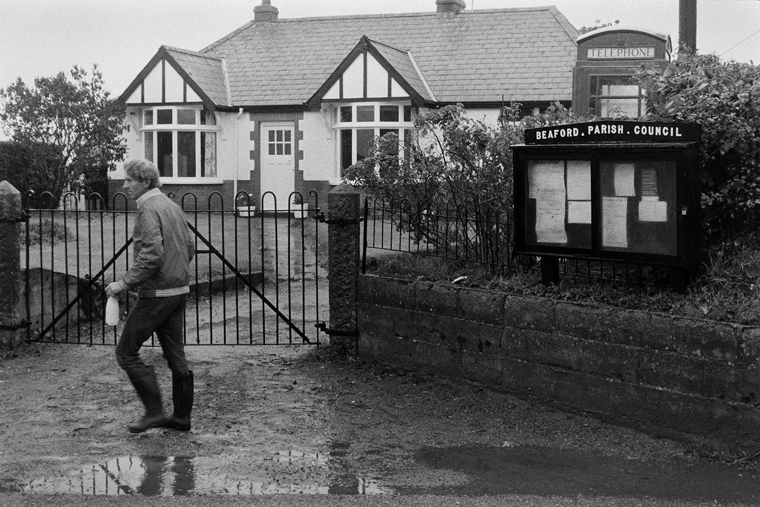 Ivor Bourne delivering milk bottles to a house in Beaford. A telephone box and Beaford Parish Council noticeboard are by the house.