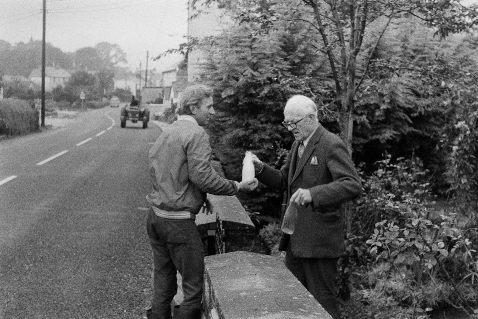Ivor Bourne handing a milk bottle to a man, over a garden wall in Beaford. The man is returning an empty bottle to Ivor. A tractor can be seen further along the street in the background.