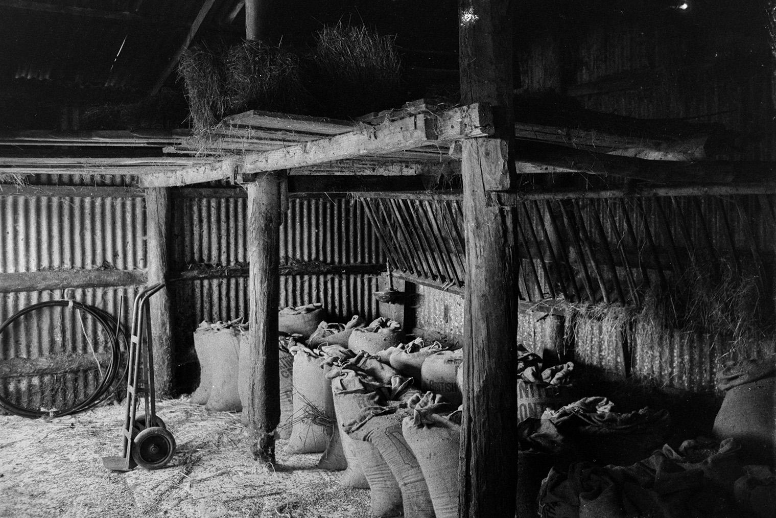 A sack truck and sacks, possibly filled with grain, in a corrugated iron barn at Mill Road Farm, Beaford. Hay bales can be seen in the tallet of the barn. The farm was also known as Jeffrys.