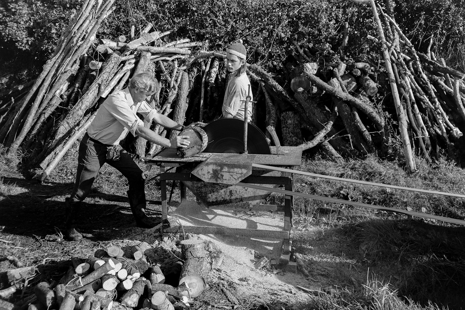 Derek Bright, on the right, helping Ivor Bourne saw logs with a circular saw, in a field at Mill Road Farm, Beaford. A log pile is resting against the hedge in the background, waiting to be sawn.  The farm was also known as Jeffrys.