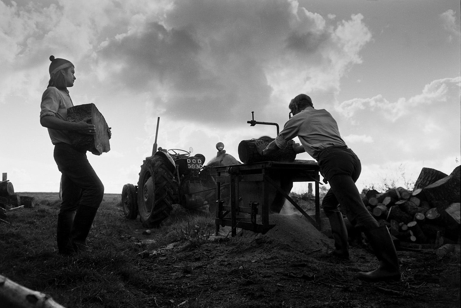 Derek Bright, on the left, helping Ivor Bourne saw logs with a circular saw, in a field at Mill Road Farm, Beaford. They are using a tractor to drive the saw. The farm was also known as Jeffrys.