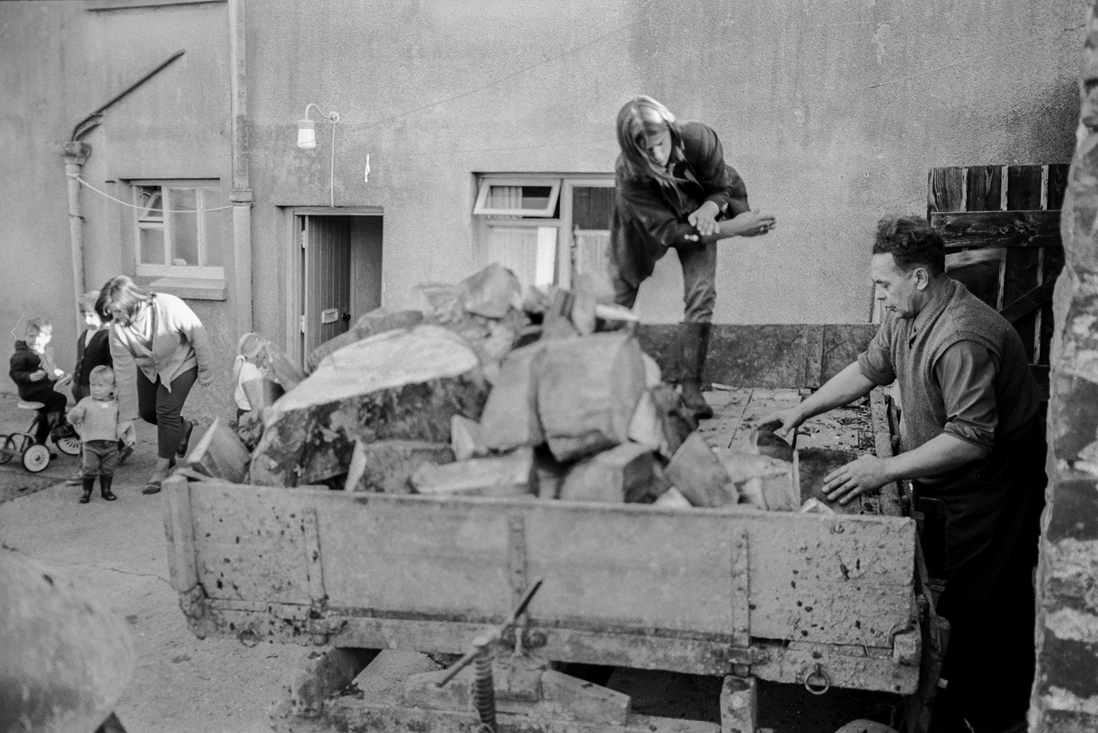 Derek Bright helping a man unload a log delivery from a trailer, possibly to a house in Beaford. A woman and children are in the background.