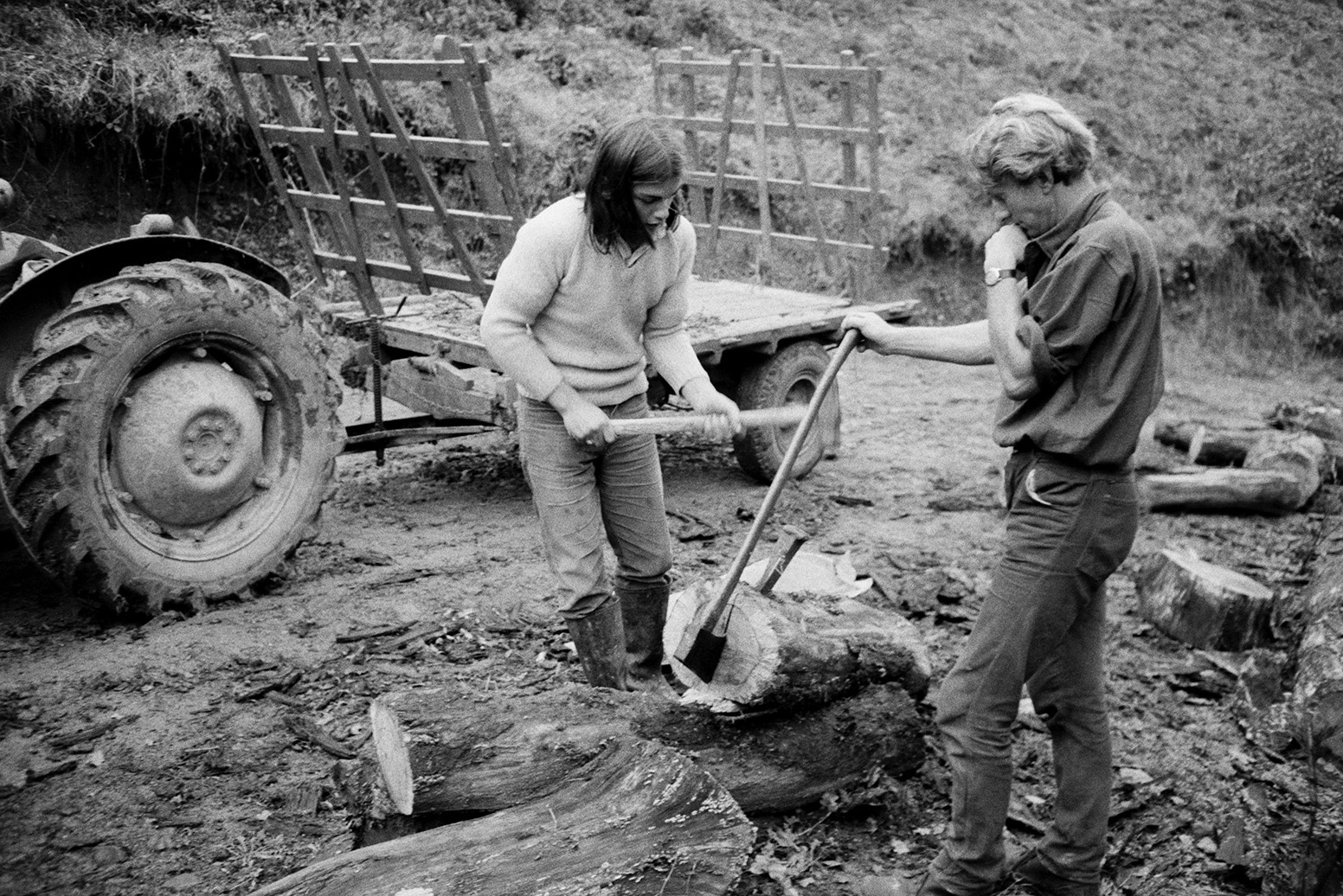 Derek Bright, on the left, helping Ivor Bourne split logs using an axe, wedges and hammer, at Mill Road Farm, Beaford. A tractor and trailer can be seen in the background. The farm was also known as Jeffrys.