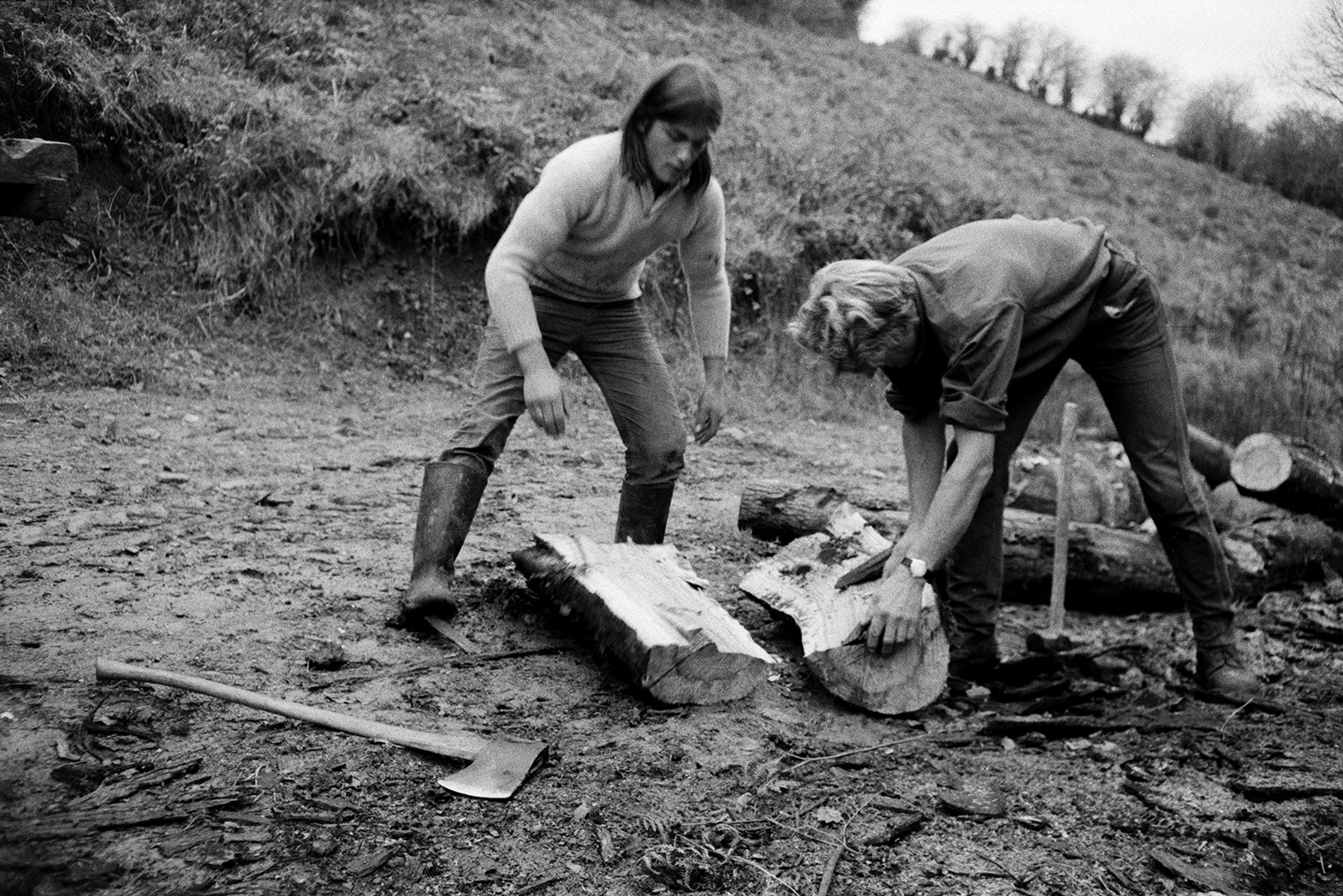 Derek Bright, on the left, helping Ivor Bourne split logs using an axe, wedges and hammer, by a grassy bank, at Mill Road Farm, Beaford. The farm was also known as Jeffrys.