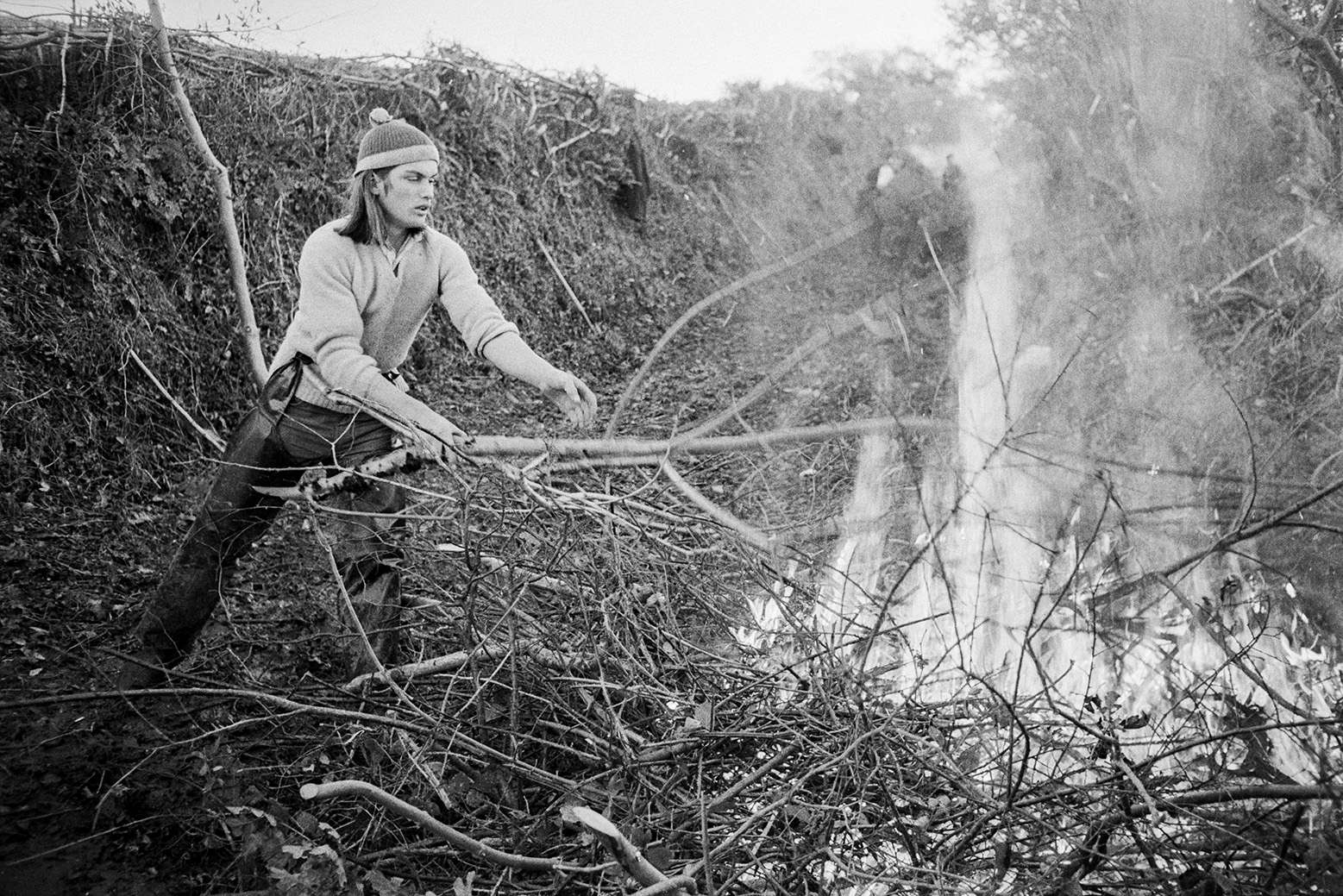 Derek Bright burning hedge cuttings, possibly in Green Lane at Beaford. He is wearing protective clothing over his legs. The hedge which has been laid can be seen in the background.