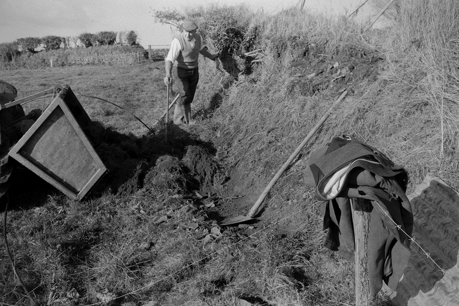 Tom Hooper clatting or building up a hedgebank with turf, in a field at Mill Road Farm, Beaford. The hedge was broken by sheep. The turf is being unloaded from a link box in front of him. The farm was also known as Jeffrys.