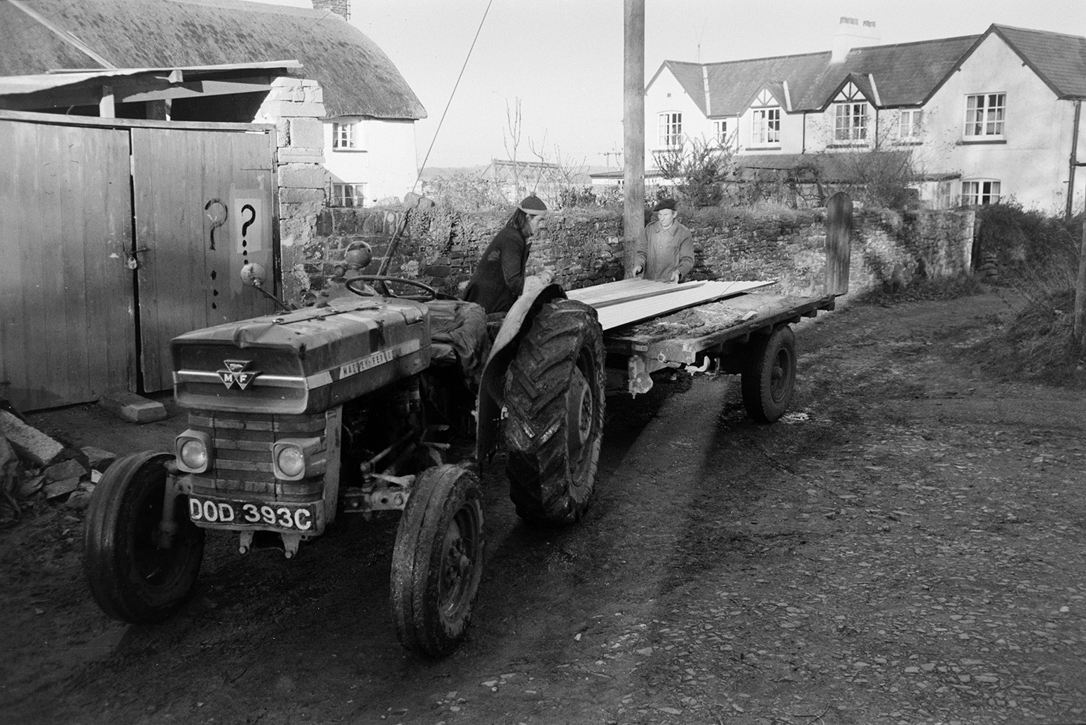 Derek Bright, in the foreground, and Ivor Bourne unloading sheets of corrugated iron from a tractor and trailer, to be used on a barn exterior, at Beaford. Houses and a thatched cottage are visible in the background. The farm was also known as Jeffrys.