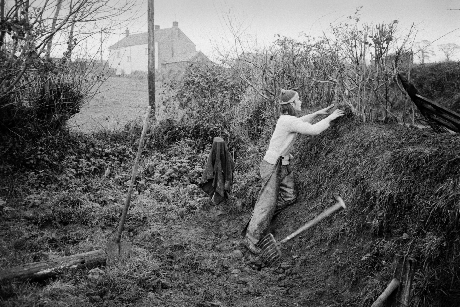 Derek Bright clatting, building up hedge bank with turf, in a field at Mill Road Farm, Beaford. A fork and spade are nearby and a farmhouse can be seen in the background. The farm was also known as Jeffrys.