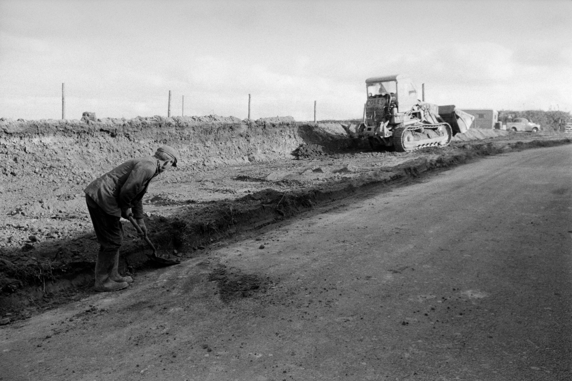 Lionel Bright clearing earth at the side of a road with a shovel, in preparation for the widening of the road. A digger is moving earth in the background.