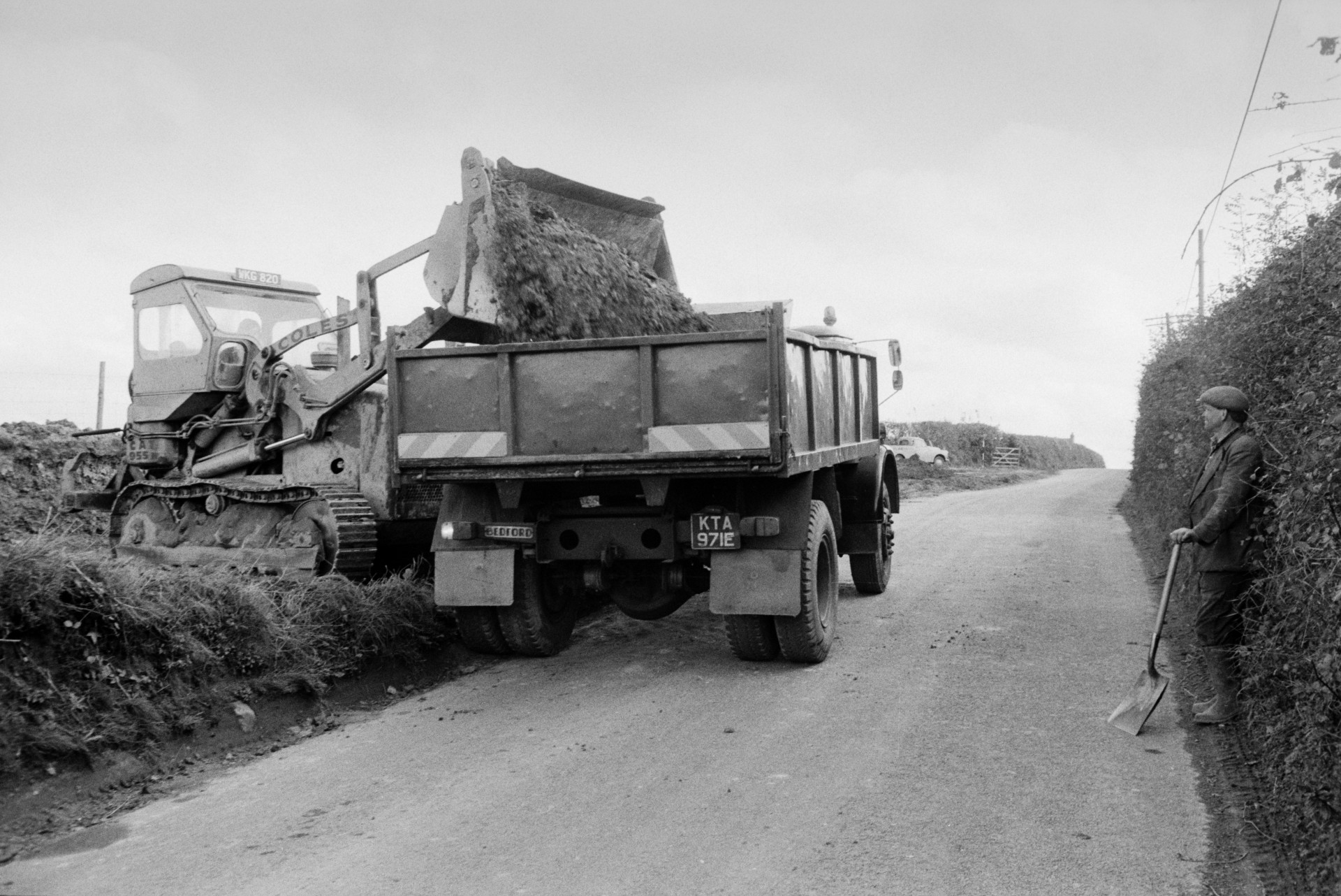 The widening of a road. A digger is loading earth into a waiting dumper truck. Lionel Bright is stood in the road holding a shovel and watching.