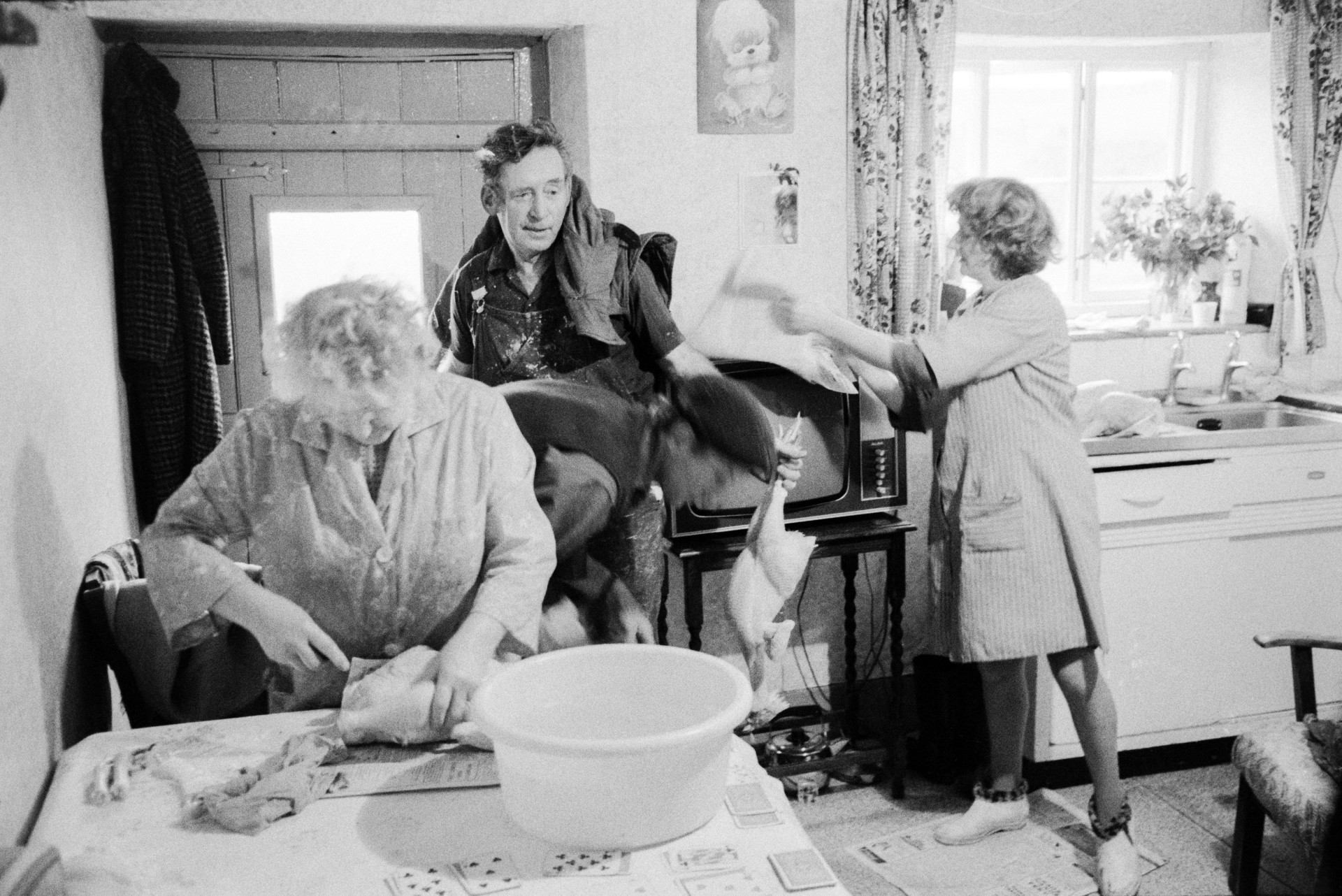 Women preparing chickens for Christmas in a kitchen at Mill Road Farm, Beaford. Ivor Bourne and Tom Spicer are also stood in the kitchen. Tom Spicer is stood by the door and holding a chicken. A television can be seen in the background. The farm was also known as Jeffrys.