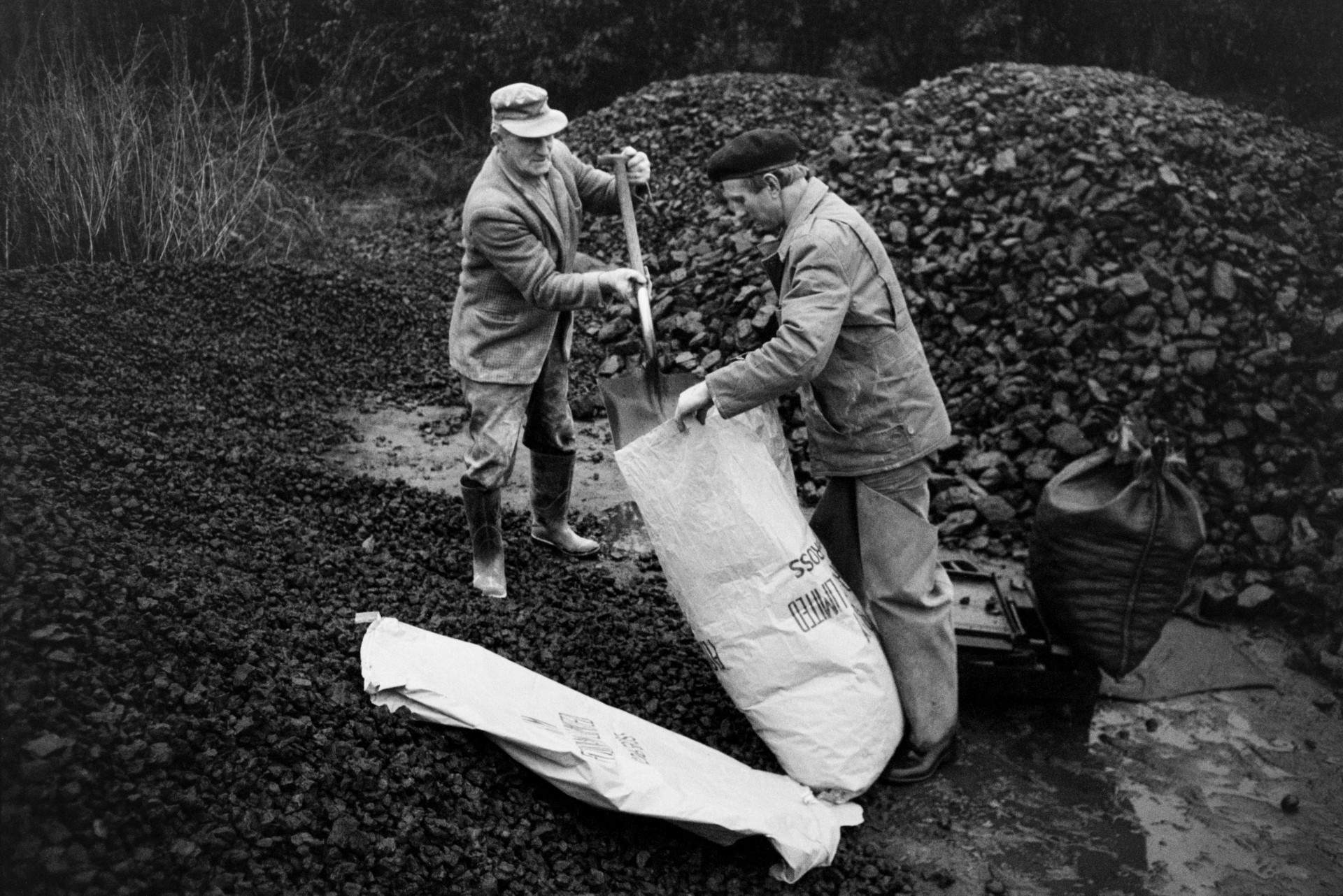 Ivor Bourne collecting coal from a coal merchant near Dolton Beacon. He is holding a sack open while another man shovels coal into it from a large mound.