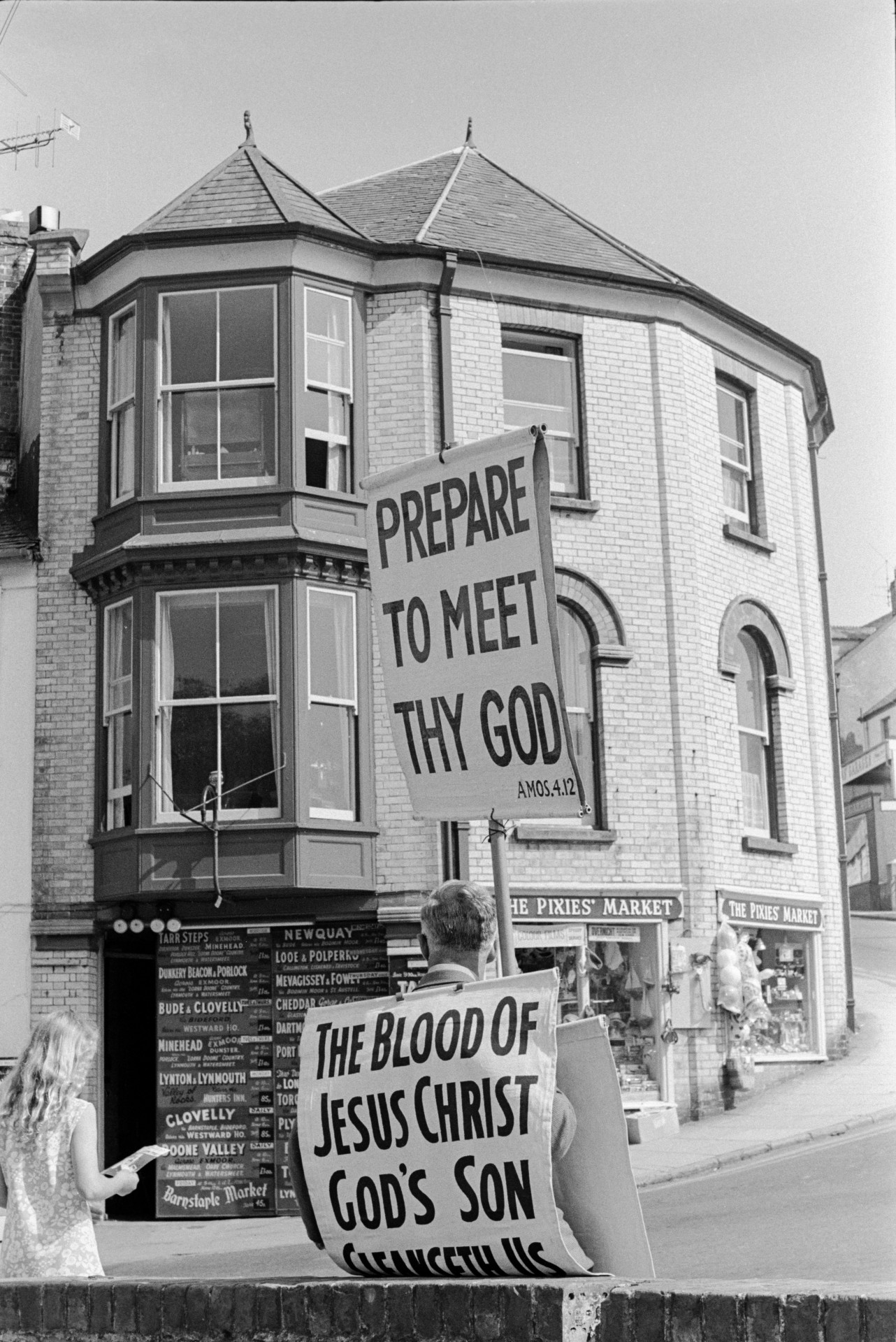 A man with religious placards sat on a wall in a street with shops, including The Pixie's Market shop, at Ilfracombe.