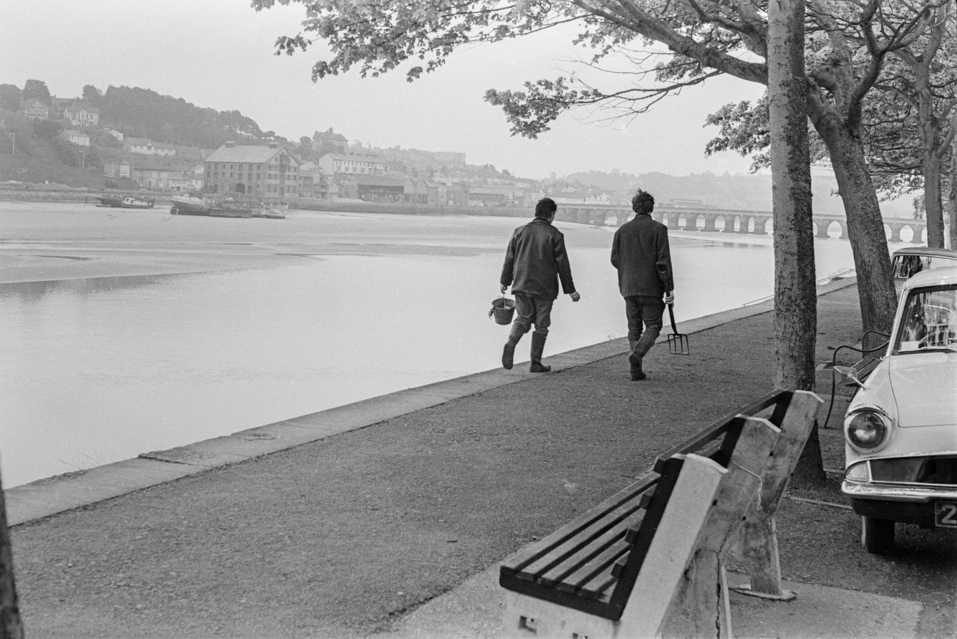 Two people walking alongside the river Torridge at Bideford. One is carrying a bucket and the other a fork. A car is parked by aa bench in the foreground. Bideford Long Bridge, also known as Bideford Old Bridge, is visible in the distance.