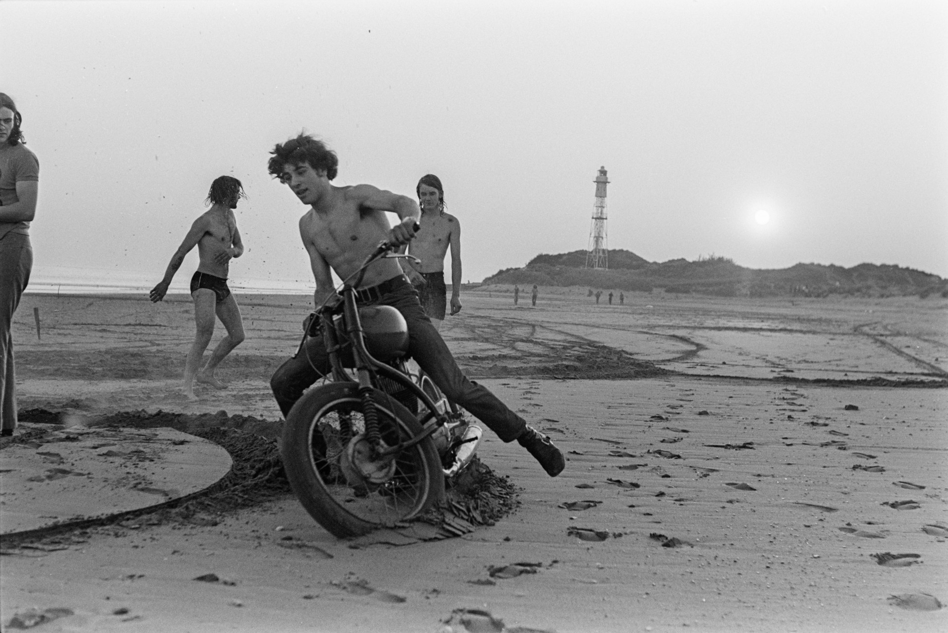 Young men running and riding a motorcycle on the beach at Braunton, at a young volunteers barbeque. A lighthouse at Crow Point is visible in the background.