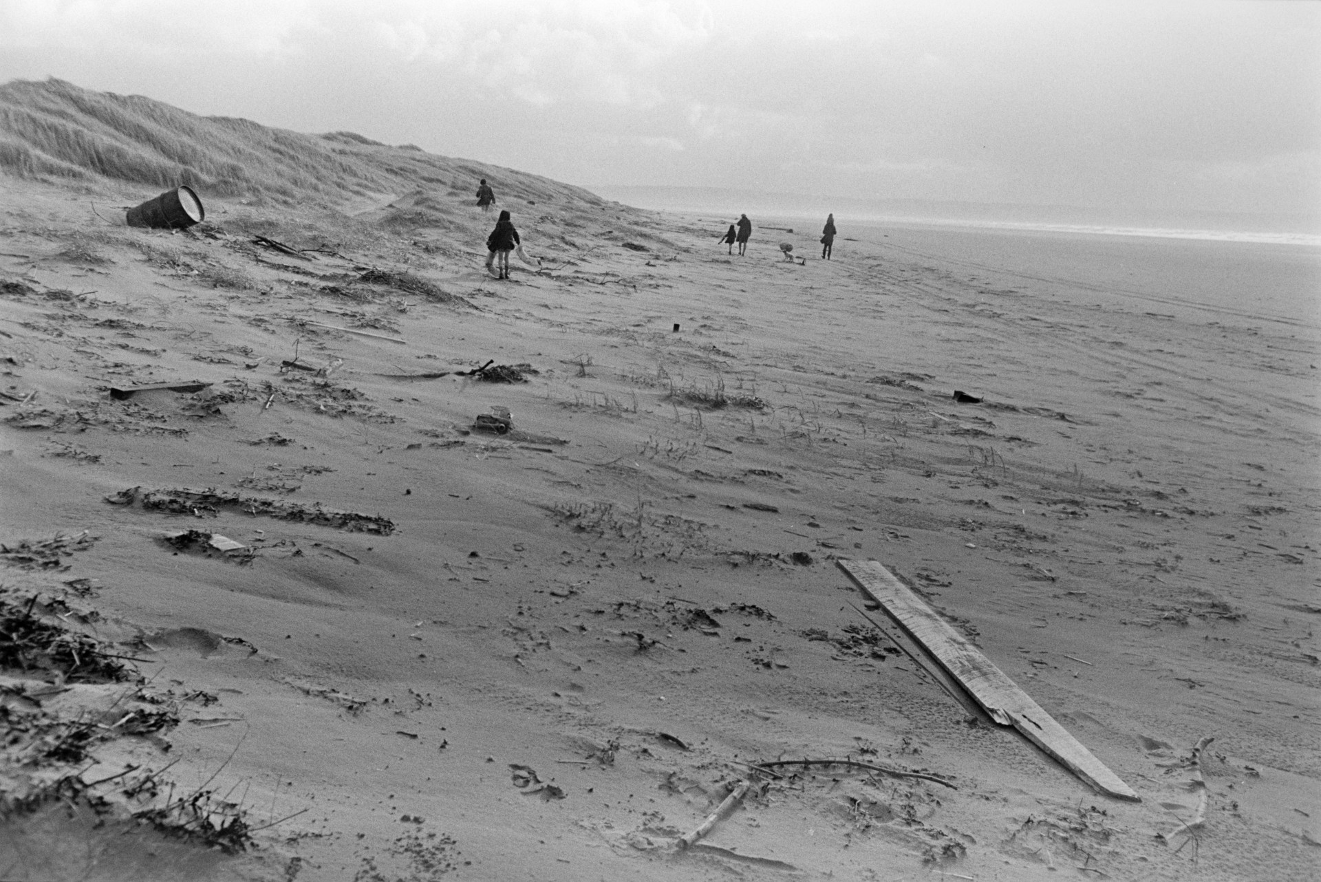 People walking along the beach at Saunton Sands, with sand dunes behind them. Debris is visible on the beach, including a barrel and a plank of wood.