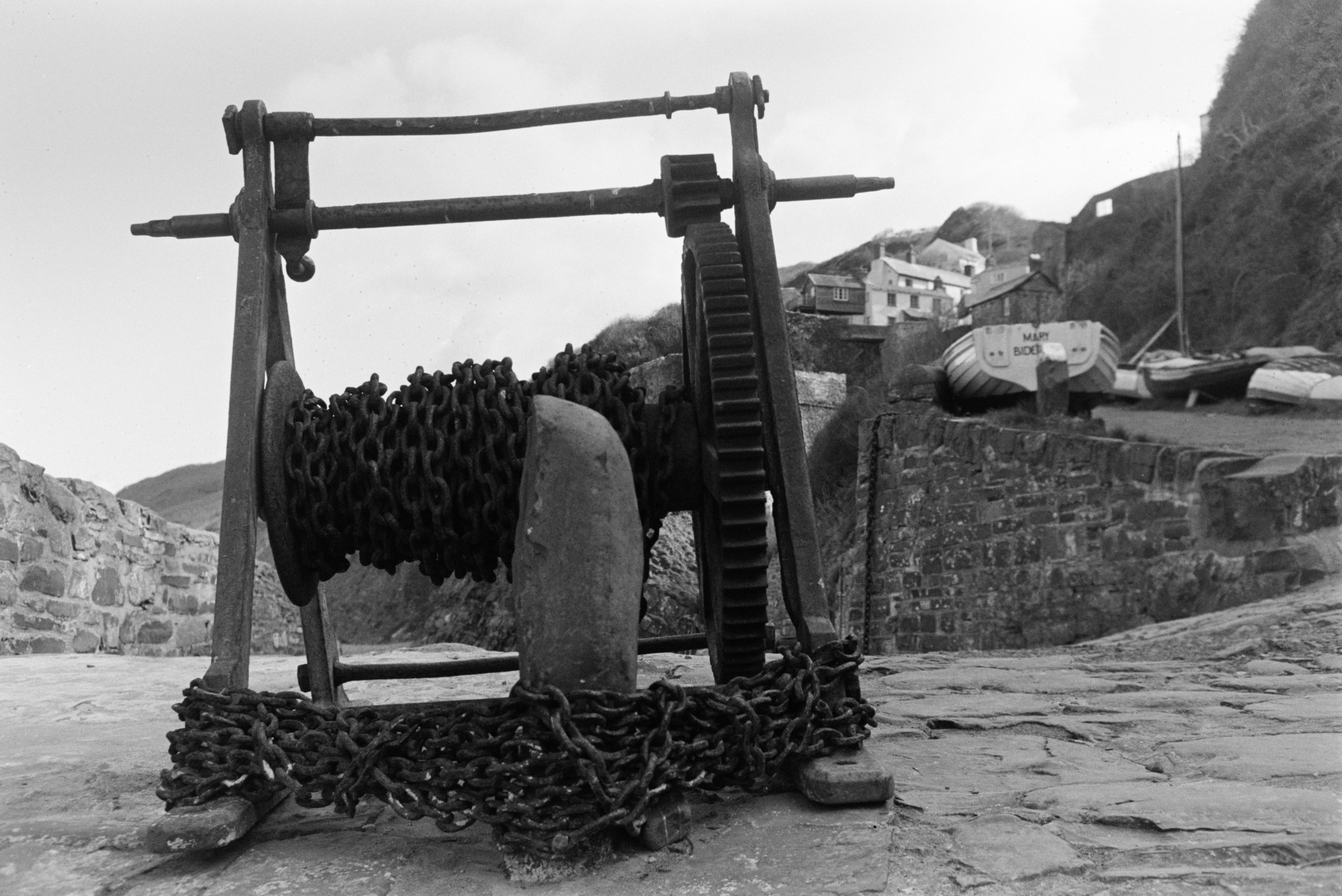Winch with chains at the top of a slipway at Bucks Mills. Boats and cottages are visible in the background.
