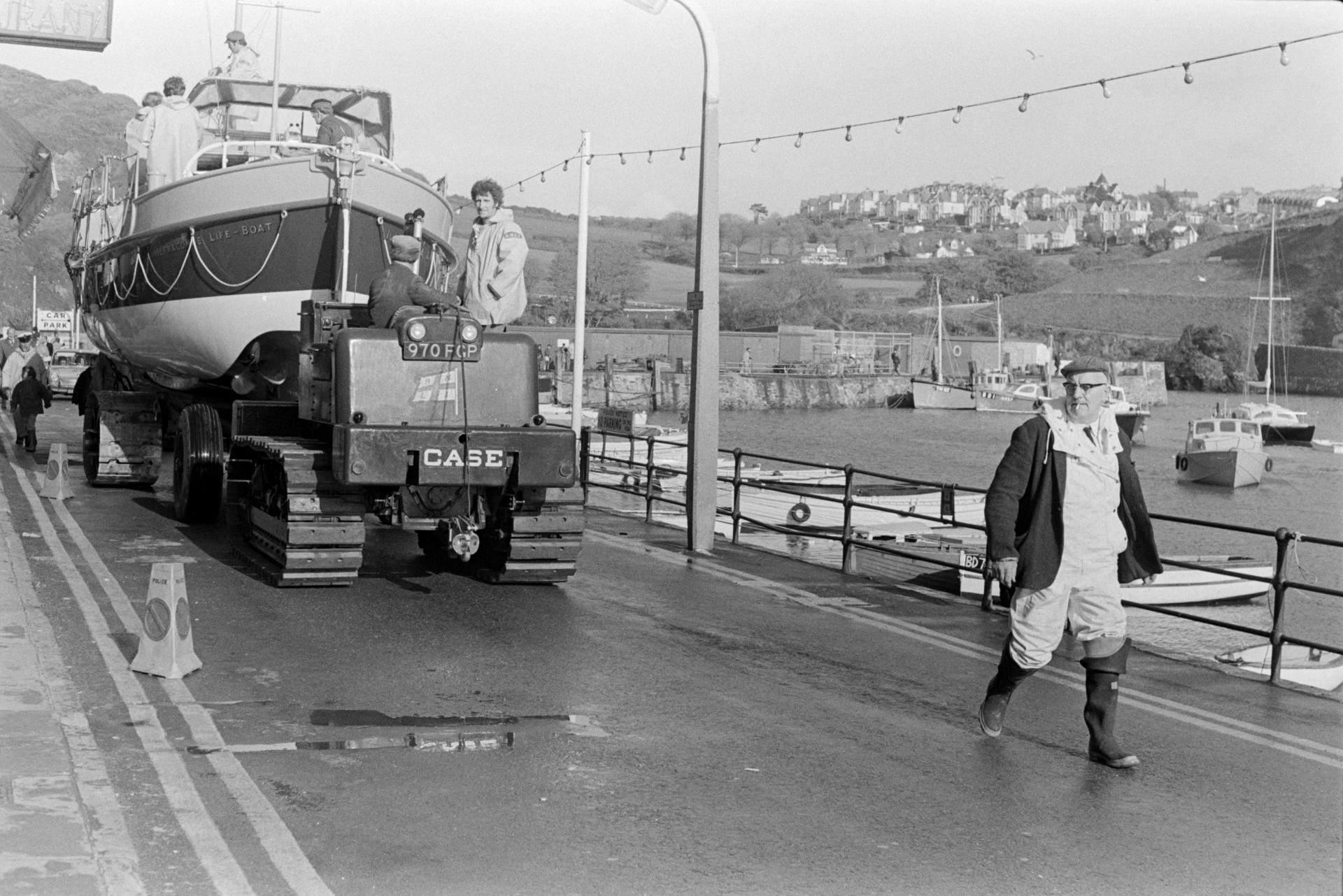 A life boat being transported through Ilfracombe on a vehicle with caterpillar tracks. It is passing the harbour. Boats are moored in the harbour in the background.